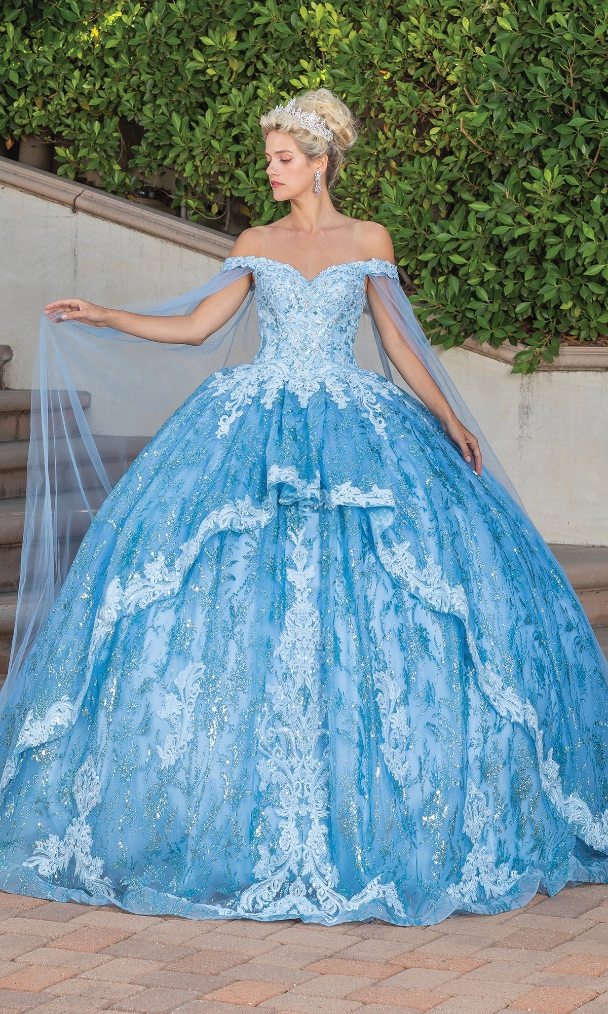 A woman stands outdoors in the Quinceanera Dress 1782 By Dancing Queen, an elaborate a-line ball gown with a sweetheart neckline and lace details, holding sheer blue fabric amid greenery and stairs.